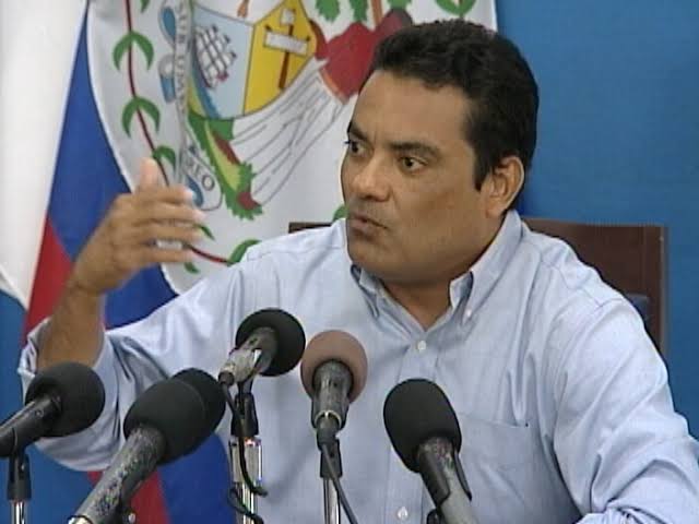 Francis Fonseca gesturing while answering questions at a press table set with several microphones, the flag of Belize behind him.