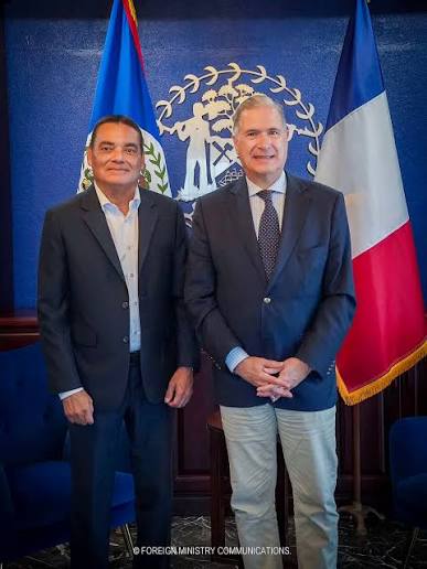 Francis Fonseca standing with a visiting representative between the flags of Belize and France.