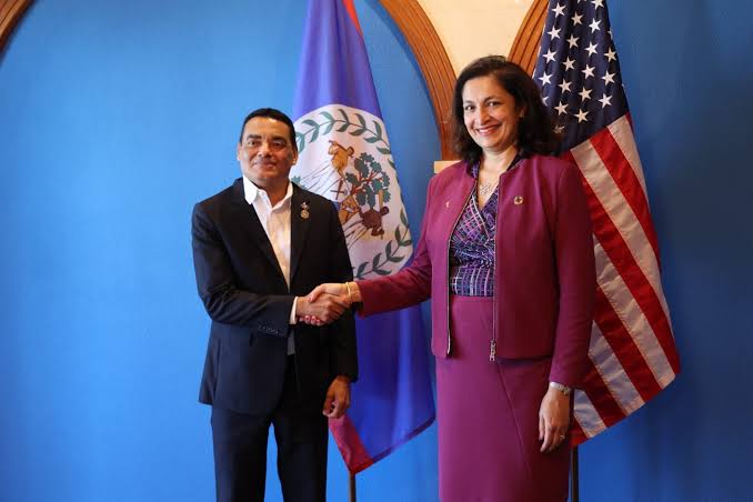Francis Fonseca shaking hands with a United States official between the flag of Belize and the flag of the United States.