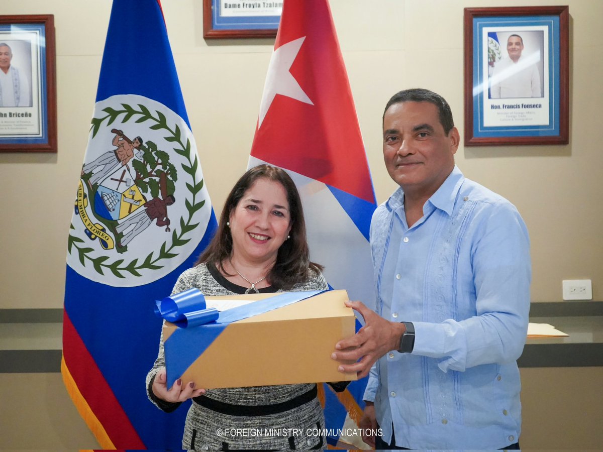 Francis Fonseca and a visiting representative jointly holding a ribboned presentation box between the flags of Belize and Cuba, framed ministerial portraits on the wall behind.