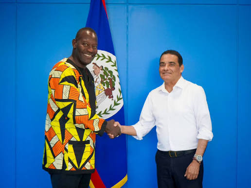 Francis Fonseca shaking hands with a visiting official wearing a patterned jacket, in front of the flag of Belize against a blue backdrop.