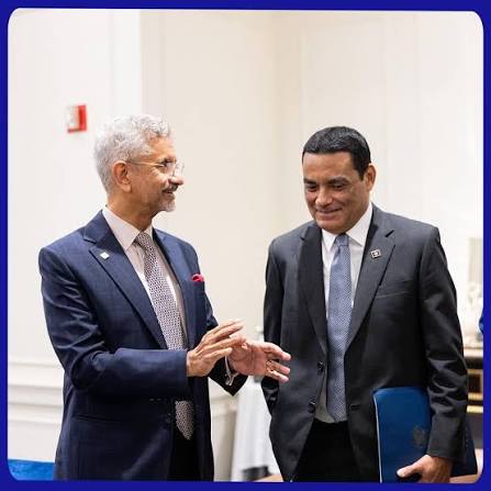 Francis Fonseca in a dark suit listening to a counterpart in conversation at a conference venue, a folder under his arm.
