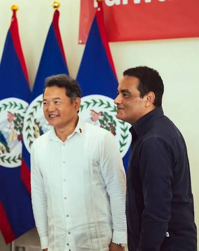 Francis Fonseca standing beside a visiting representative in front of three Belizean national flags at a reception.
