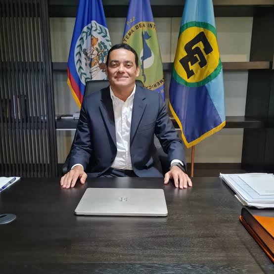 Francis Fonseca at his ministerial desk, with the flags of Belize, the Central American Integration System and the Caribbean Community behind him.