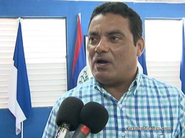 Francis Fonseca answering a broadcast interviewer's question in a party office, the flag of Belize behind him.