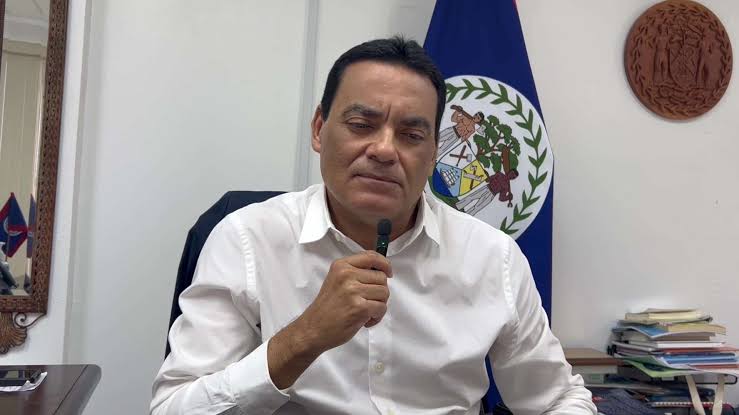 Francis Fonseca speaking into a handheld microphone at his office desk, the flag of Belize and a carved coat of arms behind him.