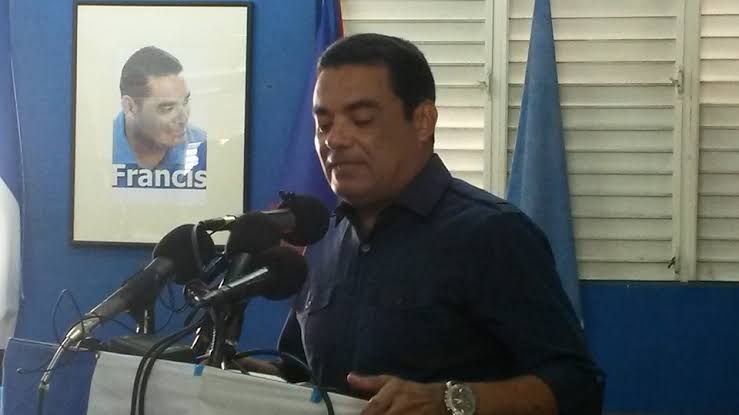 Francis Fonseca speaking from a lectern banked with microphones, a framed campaign portrait captioned Francis on the wall behind him.