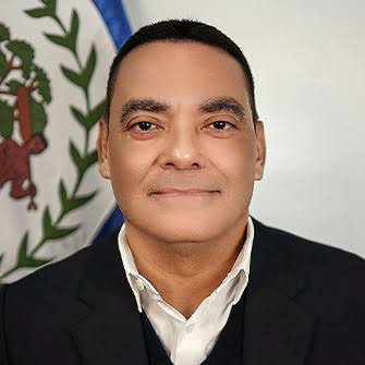 Portrait of Francis Fonseca in a dark jacket in front of the coat of arms on the flag of Belize.