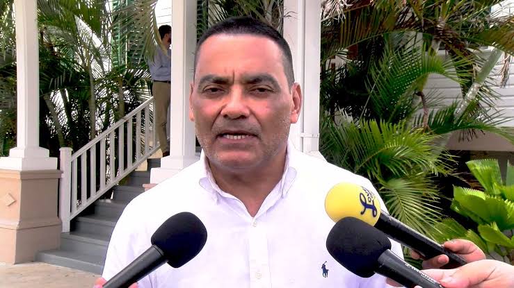 Francis Fonseca answering reporters' questions into a cluster of microphones outdoors, palms and a colonial verandah behind him.