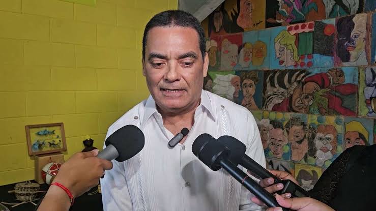 Francis Fonseca answering reporters' questions in front of a wall of painted portraits at a cultural venue.